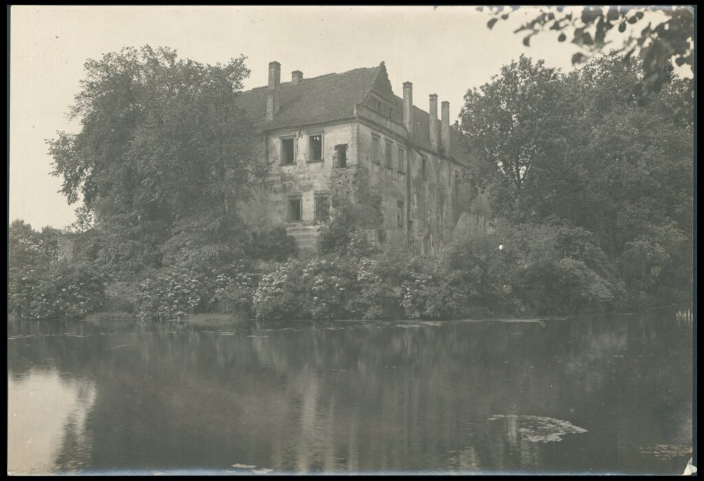 Czarno-białe zdjęcie zniszczonego budynku otoczonego drzewami. Na pierwszym planie woda.