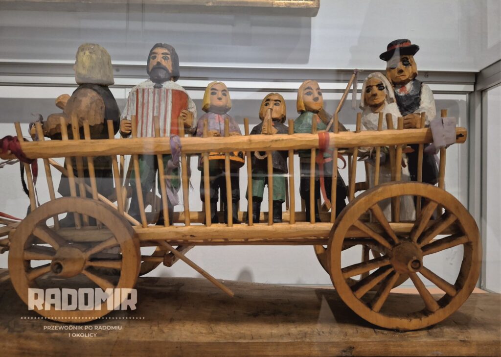 Zdjęcie drewnianej, realistycznej rzeźby. Przedstawia wóz, na którym stoją postacie z instrumentami muzycznymi oraz para młoda. 