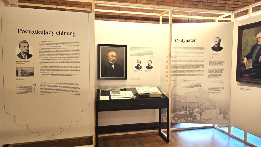 Zdjęcie. Białe ścianki z czarnymi napisami. W środkowej części portret mężczyzny ubranego na czarno. Poniżej czarny stolik od góry przykryty szklanym kloszem. W środku książki i dokumenty.